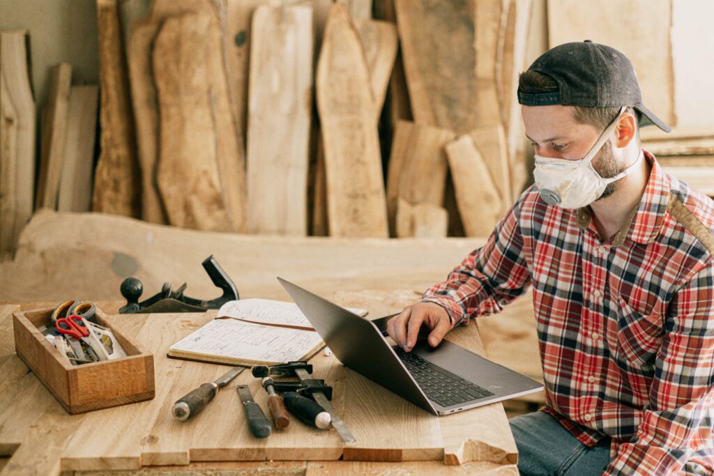 A man wearing a mask is working on a laptop next to tools in a workshop.